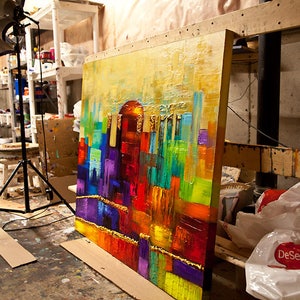 May include: A large abstract painting with a colorful cityscape scene. The painting is on a wooden easel and is leaning against a wall. The painting is done in a variety of colors, including red, orange, yellow, green, blue, and purple.