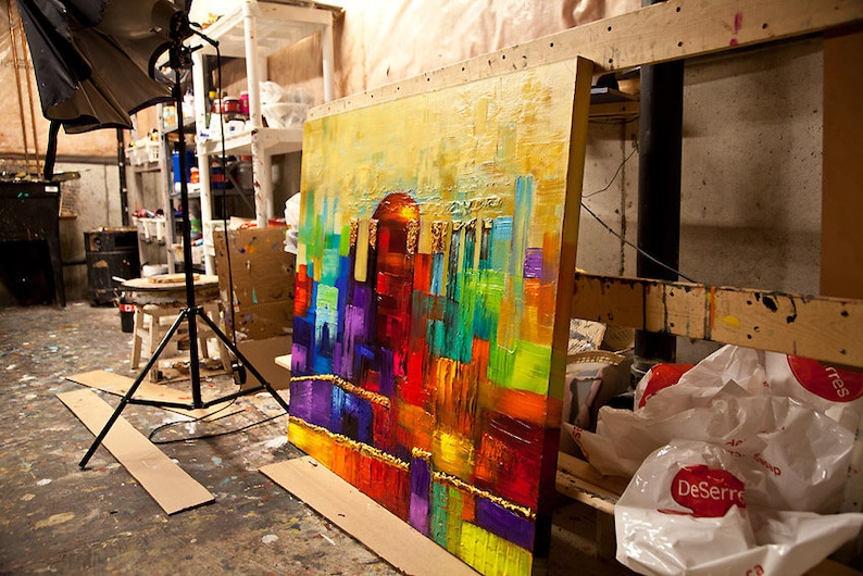 May include: A large abstract painting with a colorful cityscape scene. The painting is on a wooden easel and is leaning against a wall. The painting is done in a variety of colors, including red, orange, yellow, green, blue, and purple.