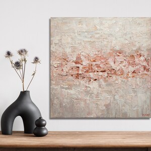 May include: Abstract painting with a textured, layered look. The painting features a light pink and white color scheme with a subtle hint of gray. The painting is framed and hanging on a white wall.