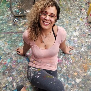 May include: A woman with curly blonde hair wearing glasses and a pink shirt sits on a floor covered in paint splatters. The text "Meet Your Artist" is at the top of the image.
