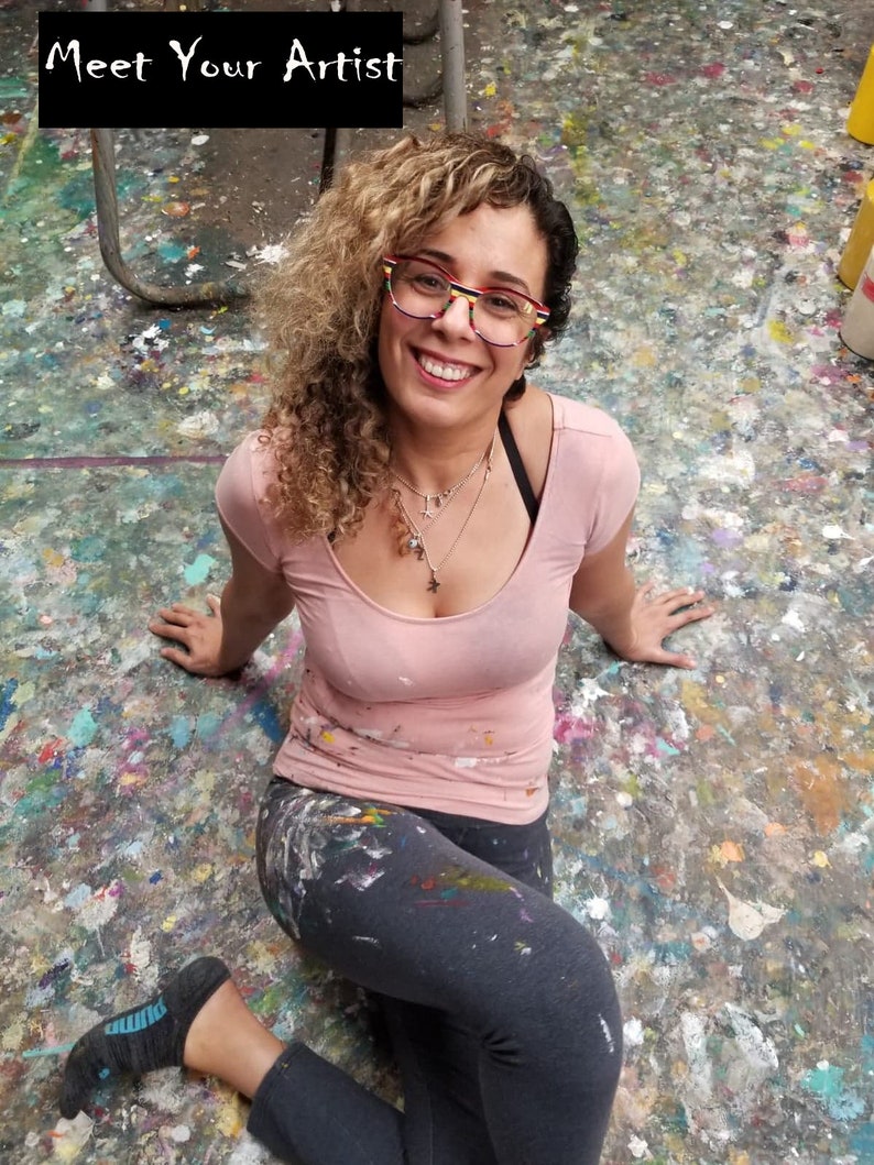 May include: A woman with curly blonde hair wearing glasses and a pink shirt sits on a floor covered in paint splatters. The text "Meet Your Artist" is at the top of the image.