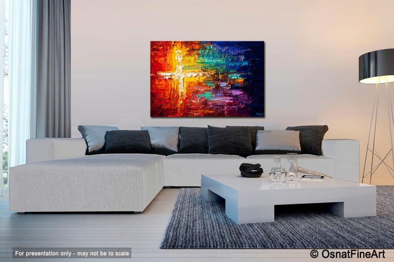 May include: Abstract painting with a white cross shape in the center, surrounded by vibrant colors like red, orange, yellow, green, blue, and purple. The painting is displayed on a white wall above a white sectional sofa with gray pillows and a white coffee table.