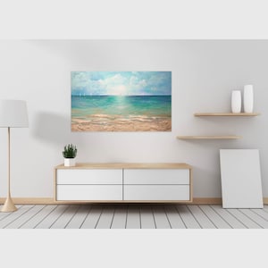 May include: A painting of a seascape with a sandy beach, blue water, and a cloudy sky. The painting is framed and hanging on a white wall.