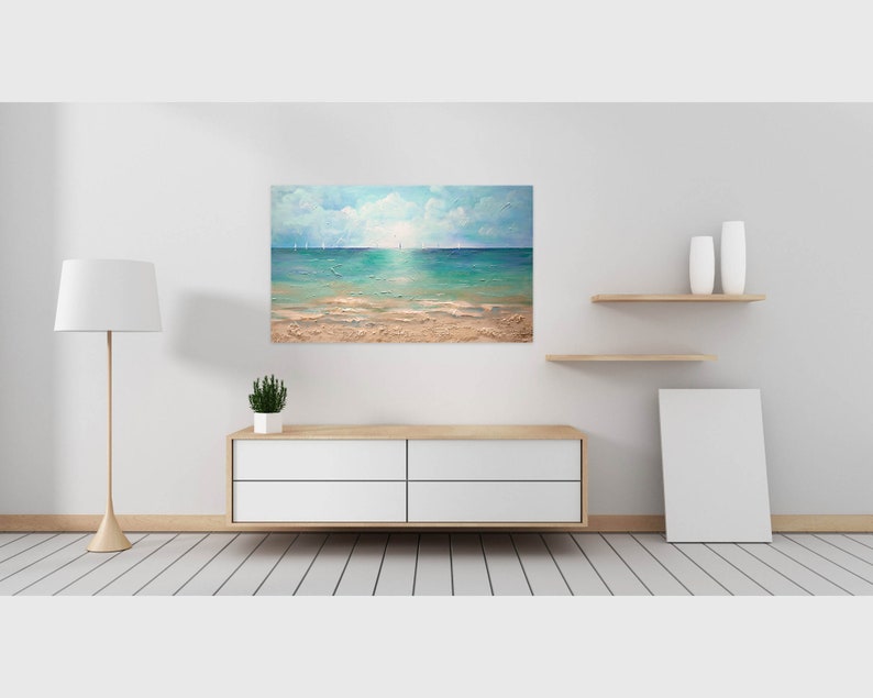 May include: A painting of a seascape with a sandy beach, blue water, and a cloudy sky. The painting is framed and hanging on a white wall.