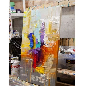 May include: Abstract painting on canvas with thick layers of paint in shades of yellow, orange, red, purple, blue, and green. The painting is leaning against a wall in an art studio.