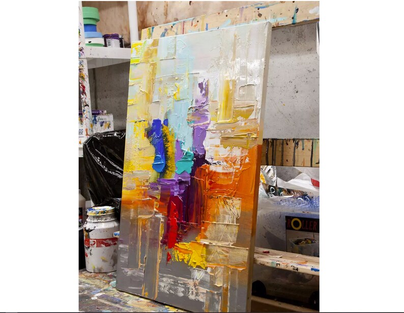 May include: Abstract painting on canvas with thick layers of paint in shades of yellow, orange, red, purple, blue, and green. The painting is leaning against a wall in an art studio.
