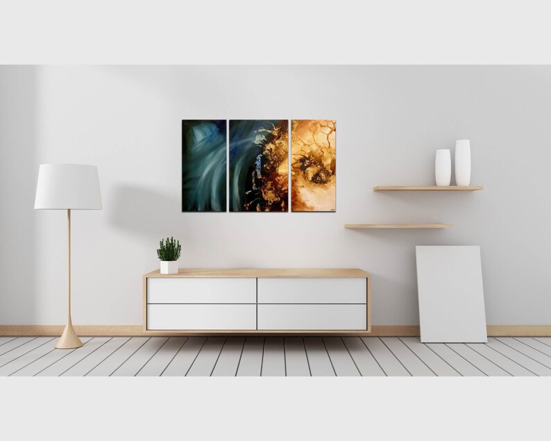 May include: Three abstract paintings in a modern living room setting. The paintings are in shades of blue, gold, and brown. The paintings are hanging on a white wall above a white dresser with four drawers. The dresser is sitting on a light wood floor. There is a white lamp on the left side of the image and a white frame on the right side of the image.