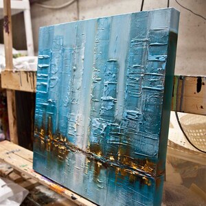 May include: Abstract painting on canvas with a textured surface. The painting features a cityscape with tall buildings in shades of blue, white, and gold.