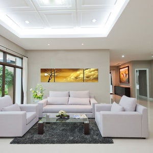 May include: A living room interior with white walls and a white ceiling. There are three white sofas arranged in a U-shape around a glass coffee table. A large abstract painting of a sunset with clouds hangs on the wall above the sofa. The room has a large window with white curtains and a view of a green garden.