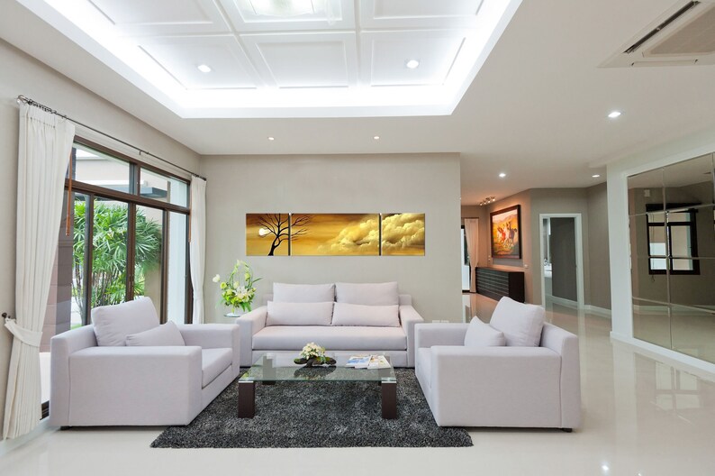 May include: A living room interior with white walls and a white ceiling. There are three white sofas arranged in a U-shape around a glass coffee table. A large abstract painting of a sunset with clouds hangs on the wall above the sofa. The room has a large window with white curtains and a view of a green garden.