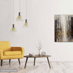 May include: Abstract painting with gold, silver, and white hues depicting a cityscape. The painting is hanging on a white wall in a living room setting with a yellow armchair and a coffee table.