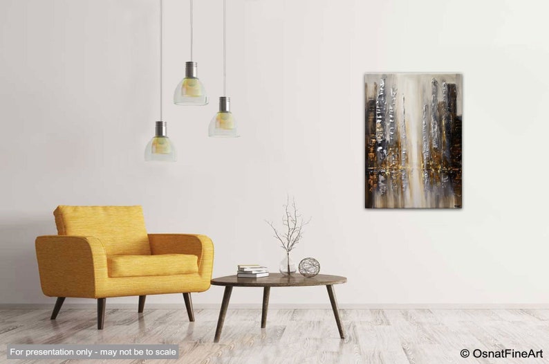 May include: Abstract painting with gold, silver, and white hues depicting a cityscape. The painting is hanging on a white wall in a living room setting with a yellow armchair and a coffee table.