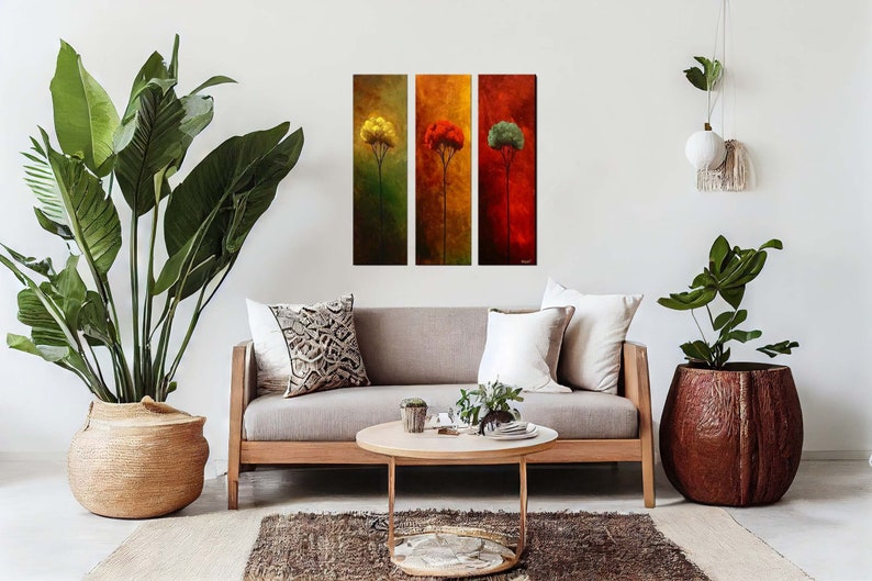 May include: Three abstract paintings of trees in different colors, yellow, red, and green, hanging on a white wall above a light brown sofa with white pillows and a coffee table with a vase of greenery.