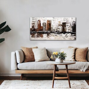 May include: A modern abstract painting of a cityscape in shades of brown, white, and black. The painting is displayed on a wall above a light gray sofa with brown and white throw pillows. The sofa is in a living room setting with a wooden coffee table and potted plants.