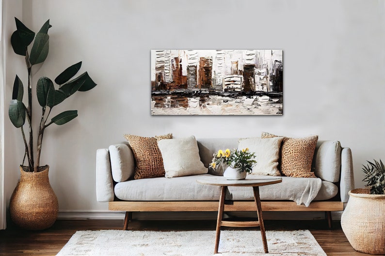 May include: A modern abstract painting of a cityscape in shades of brown, white, and black. The painting is displayed on a wall above a light gray sofa with brown and white throw pillows. The sofa is in a living room setting with a wooden coffee table and potted plants.