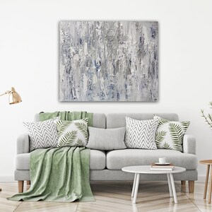 May include: A gray and white abstract painting hangs above a gray sofa with green throw pillows and a green throw blanket. The sofa is in a living room with a gold floor lamp and a small wooden side table with a vase of greenery.