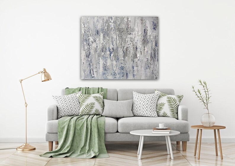 May include: A gray and white abstract painting hangs above a gray sofa with green throw pillows and a green throw blanket. The sofa is in a living room with a gold floor lamp and a small wooden side table with a vase of greenery.