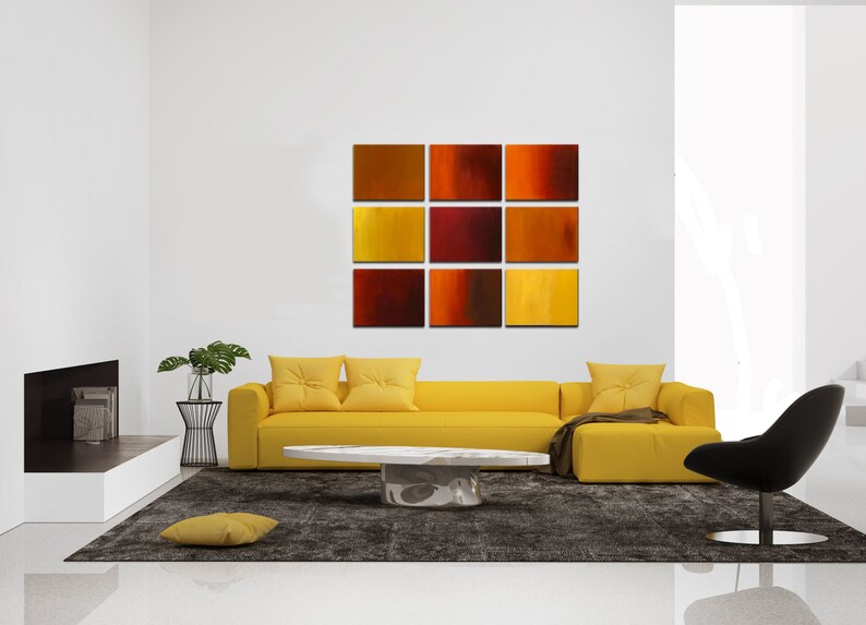 May include: A yellow sectional sofa with three pillows sits in front of a wall with a nine-piece abstract painting in shades of red, orange, and yellow. The painting is made up of nine square canvases.