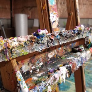 May include: A wooden artist's easel covered in dried paint. The paint is a variety of colours, including white, blue, green, yellow, and red. The easel has a shelf for holding paint tubes or brushes.