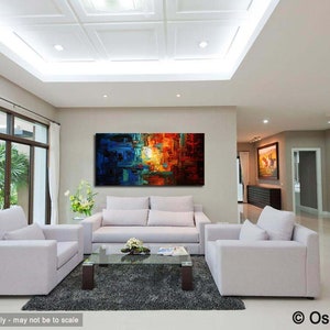 May include: Abstract painting with blue, orange, and yellow hues. The painting is hanging on a wall in a living room with white couches and a coffee table.