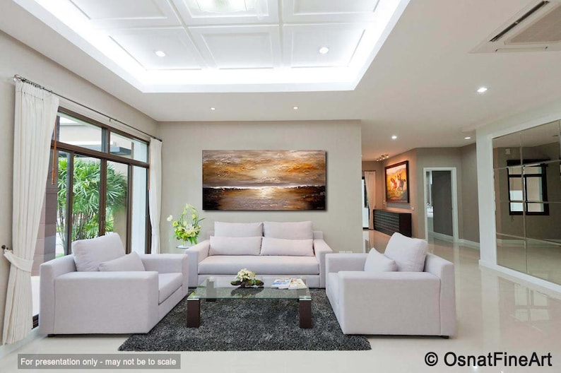 May include: A large abstract painting of a sunset over a body of water hangs on a wall in a living room. The painting is in shades of gold, brown, and white. The living room is decorated with white furniture and a gray rug.