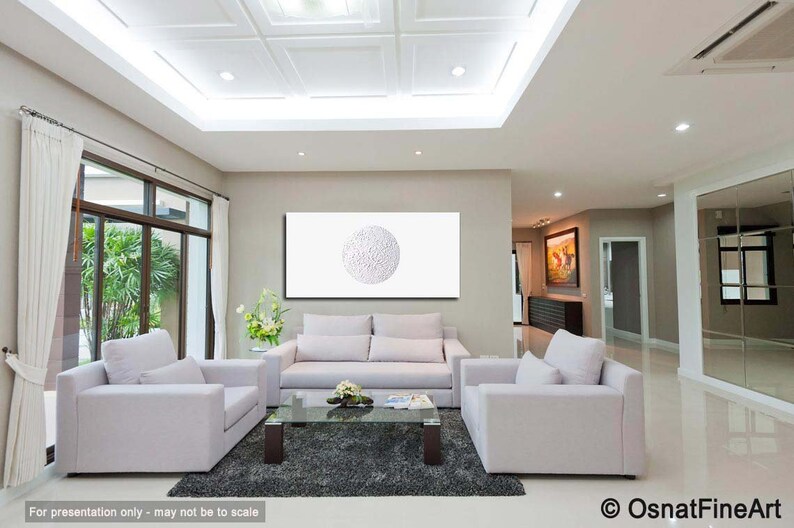 May include: A white living room with two white sofas, a coffee table, and a large white framed canvas with a white circle design. The room has a high ceiling with recessed lighting and white curtains.