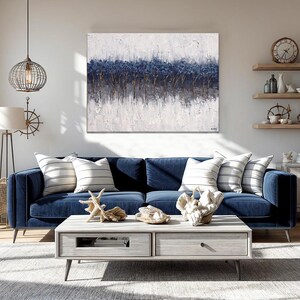 May include: A navy blue velvet sofa with striped pillows and a light gray coffee table. The room features a large abstract painting with blue and white tones, and decorative items like coral and a starfish. A coastal theme.