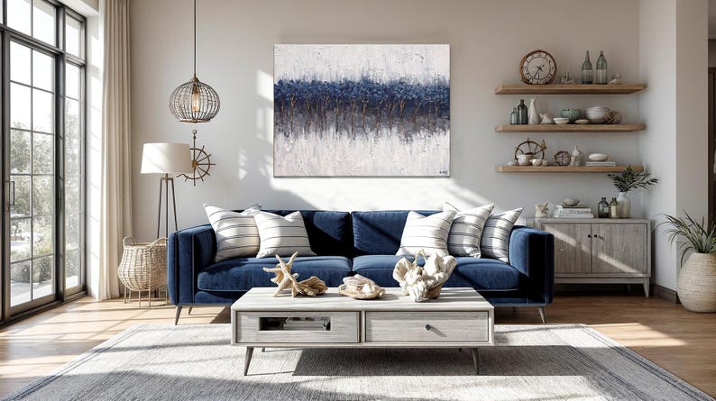 May include: A navy blue velvet sofa with striped pillows and a light gray coffee table. The room features a large abstract painting with blue and white tones, and decorative items like coral and a starfish. A coastal theme.