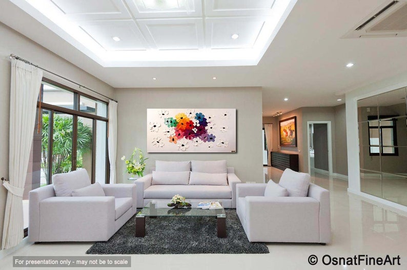 May include: A colorful abstract painting of flowers in shades of pink, red, orange, yellow, blue, and green. The painting is on a white canvas and is framed in a white frame. The painting is hanging on a wall in a living room with white couches and a coffee table.