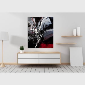 May include: Abstract painting with black, white, and red paint splatters and brushstrokes. The painting is framed and hanging on a white wall above a wooden dresser.