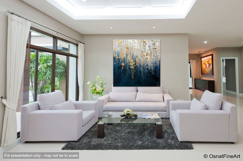 May include: An abstract painting with a textured surface, featuring a blend of deep blue and gold tones. The artwork is displayed above a white sofa in a modern living room setting. The room includes white armchairs, a glass coffee table, and a gray rug.