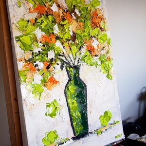 May include: An original oil painting of a bouquet of flowers in a green vase. The flowers are a mix of green, orange, and white, with thick brushstrokes creating a textured effect. The vase is a dark green color, and the background is white.