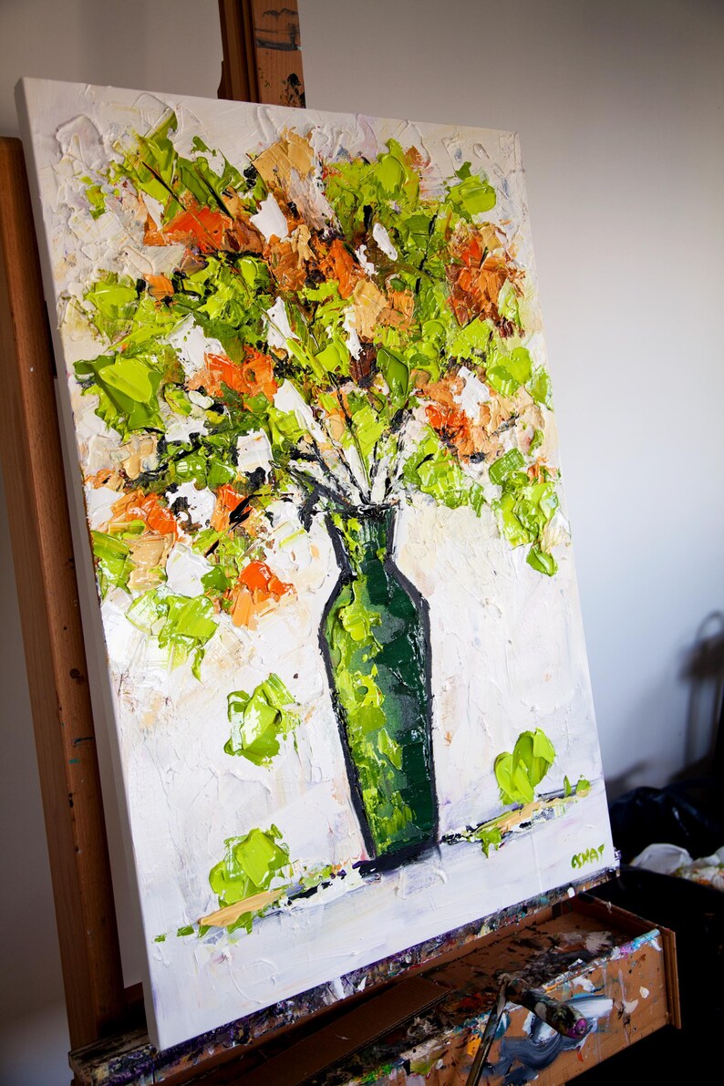 May include: An original oil painting of a bouquet of flowers in a green vase. The flowers are a mix of green, orange, and white, with thick brushstrokes creating a textured effect. The vase is a dark green color, and the background is white.