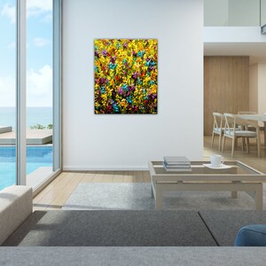 May include: Abstract painting of a field of colorful flowers in shades of yellow, blue, green, and purple. The painting is framed and hanging on a white wall in a modern living room.