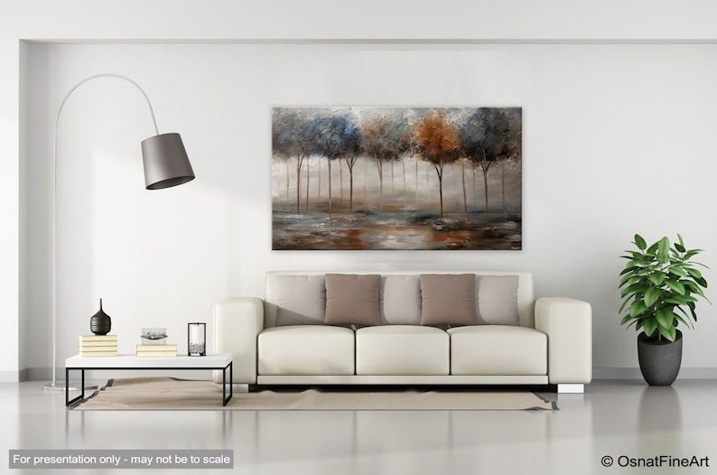 May include: A painting of a forest scene with trees in shades of blue, brown, and green. The trees are silhouetted against a misty background. The painting is displayed on a white wall above a white sofa with brown pillows.