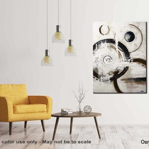 May include: Abstract painting with black and white circles and lines on a white background. The painting has a brown and white sun with Roman numerals in the center. The painting is hanging on a white wall in a living room setting.
