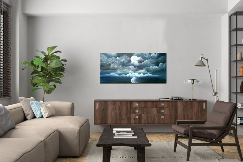 May include: A painting of a full moon in a dark blue sky with white clouds. The moon is surrounded by clouds and the sky is dark blue. The painting is framed and hanging on a wall in a living room.