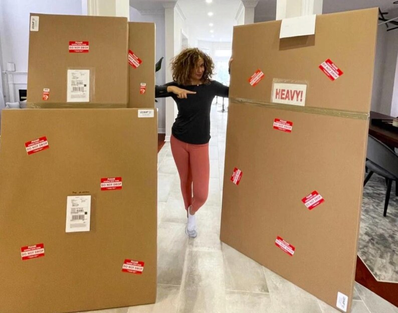 May include: A woman stands in front of several large cardboard boxes. The boxes are brown and have red warning labels. One box is labeled "HEAVY!"