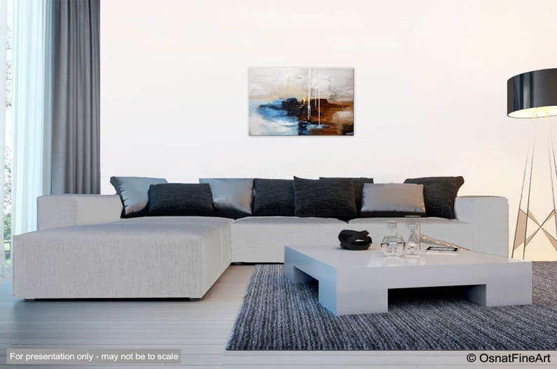 May include: Abstract painting with shades of blue, brown, and white. The painting is hanging on a white wall above a gray sectional sofa with black pillows. The sofa is in a living room setting with a gray rug and a white coffee table.