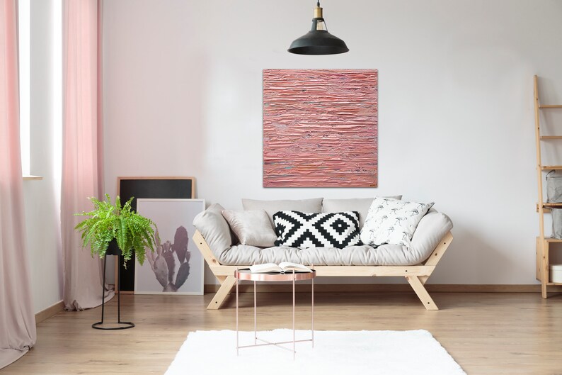 May include: A light pink abstract painting with horizontal brushstrokes hangs above a light beige sofa with black and white patterned pillows. The sofa is in a living room with a potted fern plant, a wooden ladder shelf, and a small round coffee table.