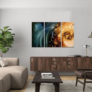 May include: A living room interior with a brown couch, a brown armchair, a coffee table, a brown cabinet, a green plant, and a three-piece abstract art piece on the wall. The art piece is made up of three canvases with a blue, green, and gold color scheme.