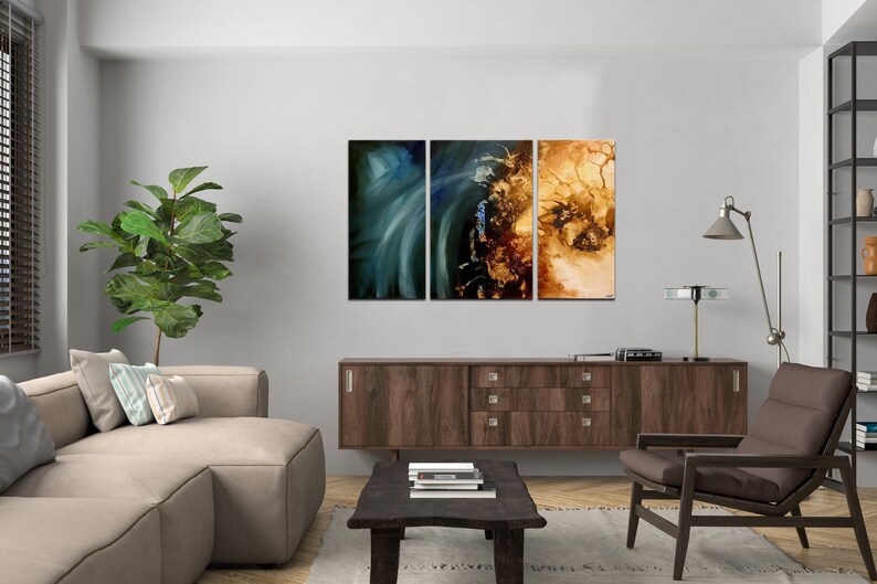 May include: A living room interior with a brown couch, a brown armchair, a coffee table, a brown cabinet, a green plant, and a three-piece abstract art piece on the wall. The art piece is made up of three canvases with a blue, green, and gold color scheme.
