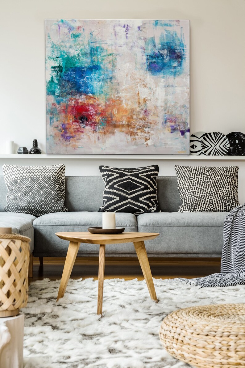May include: A gray sectional sofa with black and white patterned throw pillows, a wooden coffee table, a woven basket, and a woven ottoman. An abstract painting with blue, green, red, and orange hues hangs above the sofa.