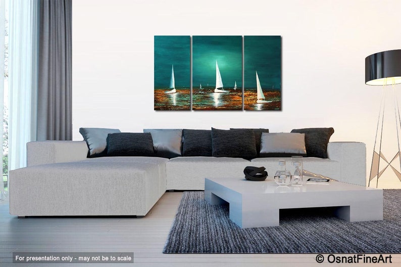 May include: A three-piece abstract painting depicting sailboats on a teal blue body of water with a golden sunset reflection. The painting is displayed on a white wall above a gray sectional sofa with black throw pillows.