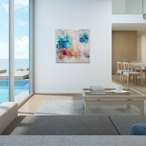 May include: Abstract painting with blue, red, and yellow colors on a white background. The painting is hanging on a white wall in a modern living room with a view of a pool and ocean.