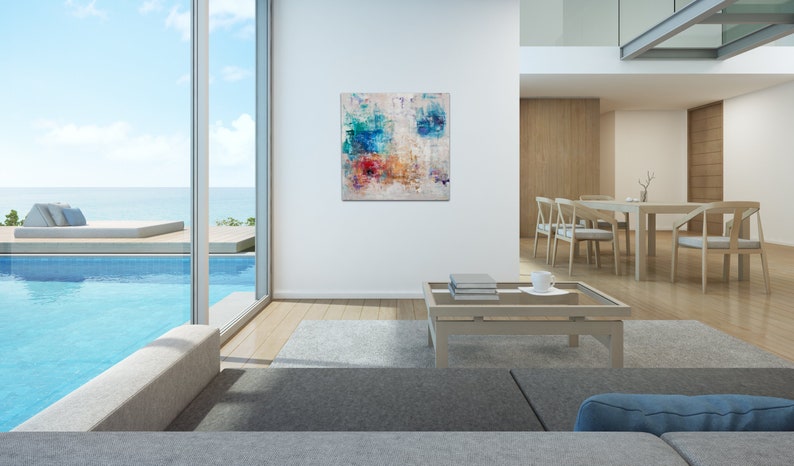 May include: Abstract painting with blue, red, and yellow colors on a white background. The painting is hanging on a white wall in a modern living room with a view of a pool and ocean.