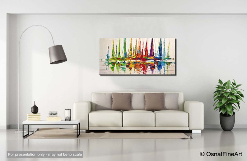 May include: Abstract painting of colorful sailboats on a white background with reflections in the water. The painting is framed and hanging on a white wall above a white couch.