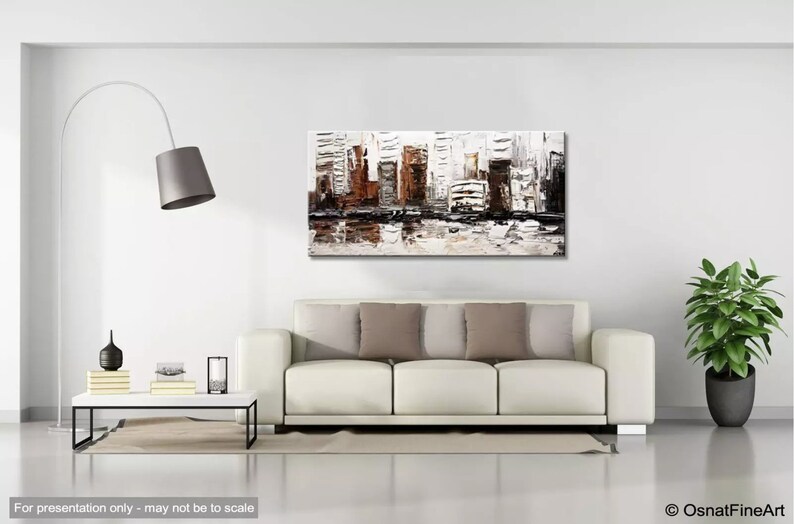 May include: Abstract painting of a city skyline in shades of brown, gray, and white. The painting is displayed on a white wall above a white sofa with brown pillows.