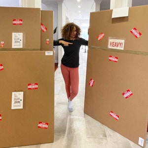 May include: A woman stands in front of four large cardboard boxes. The boxes are brown and have red warning labels that say "Please Do Not Stack". One of the boxes is labeled "Heavy!"