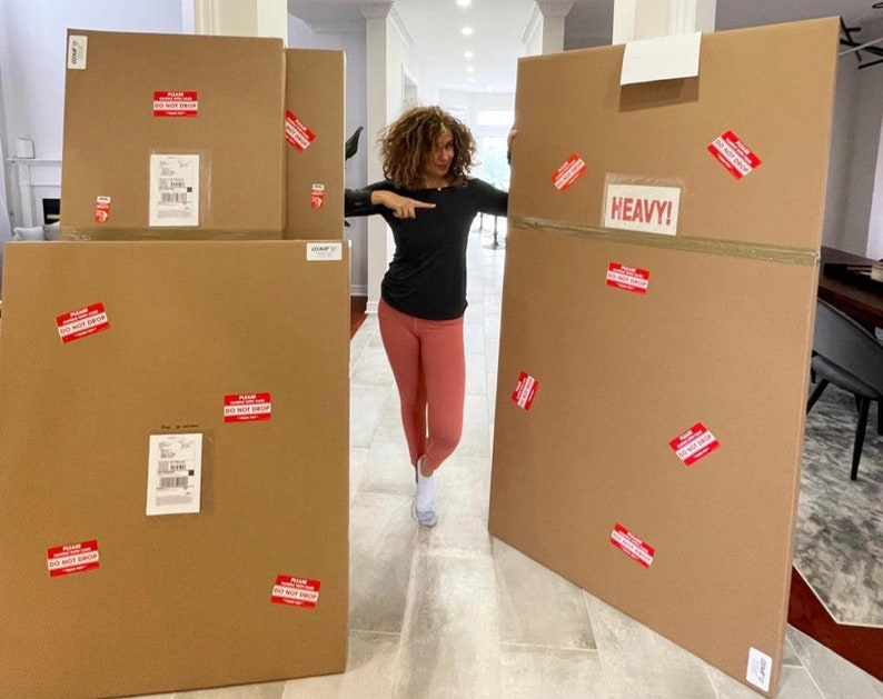 May include: A woman stands in front of four large cardboard boxes. The boxes are brown and have red warning labels that say "Please Do Not Stack". One of the boxes is labeled "Heavy!"
